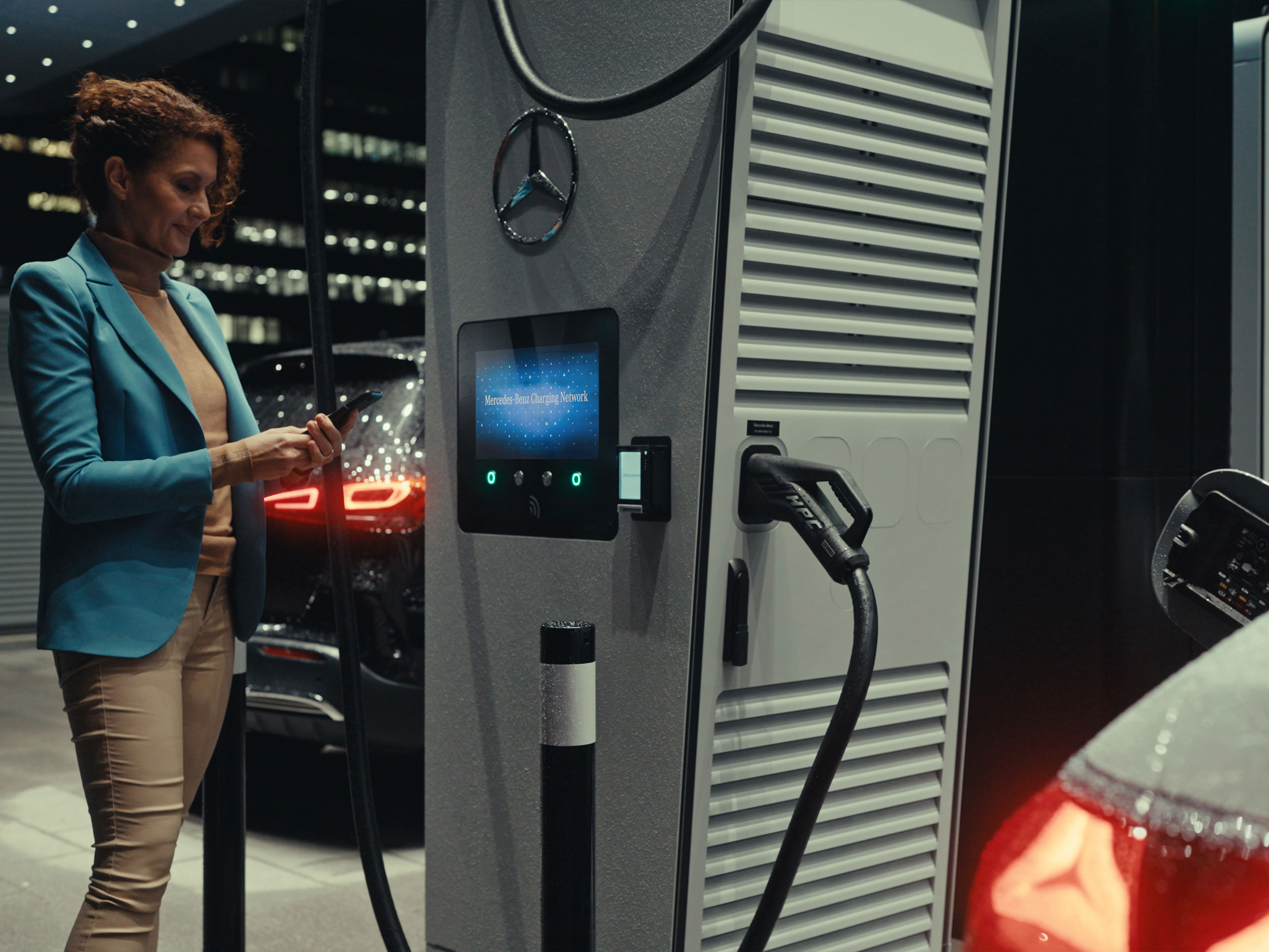 App Mercedes-Benz | Ricarica in viaggio | Mercedes-Benz Una donna che tiene in mano uno smartphone, in piedi accanto a una stazione di ricarica.