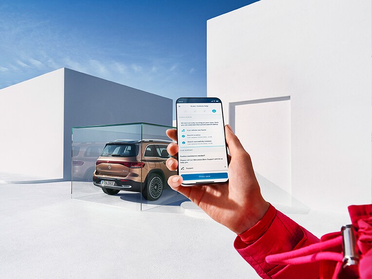 Una persona tiene un telefono cellulare e utilizza l'app Mercedes. Sullo sfondo si vede una EQB parcheggiata.