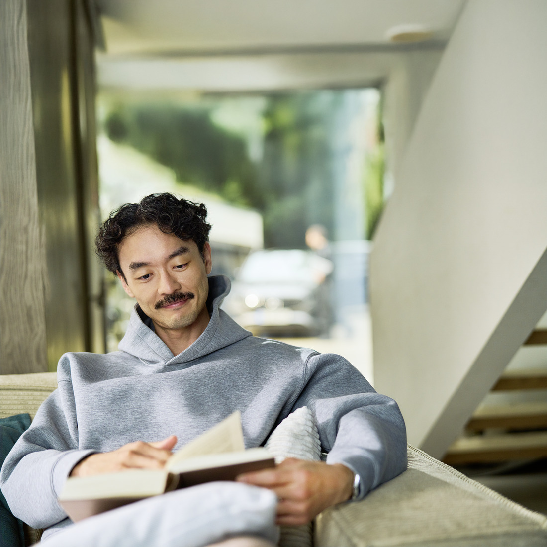 Un cliente rilassato che usufruisce del servizio assistenza mobile a casa propria Un cliente seduto in casa intento a leggere un libro. Da una finestra alle sue spalle si vede un'auto durante la manutenzione effettuata da un tecnico.