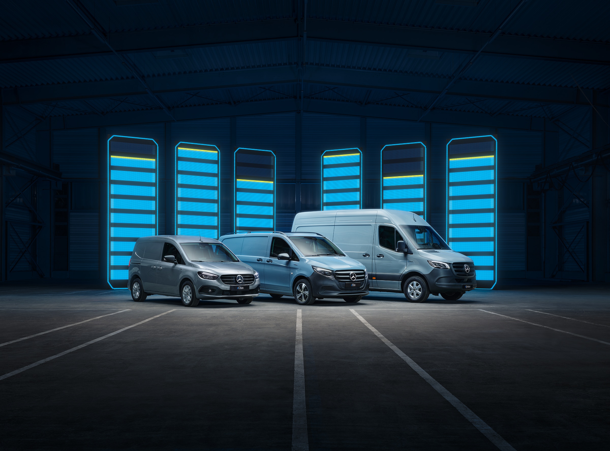 La gamma dei veicoli commerciali elettrici Mercedes-Benz | Storia dei veicoli commerciali | Mercedes-Benz La gamma dei veicoli commerciali elettrici Mercedes-Benz esposta in un magazzino futuristico con colonnine di ricarica illuminate.