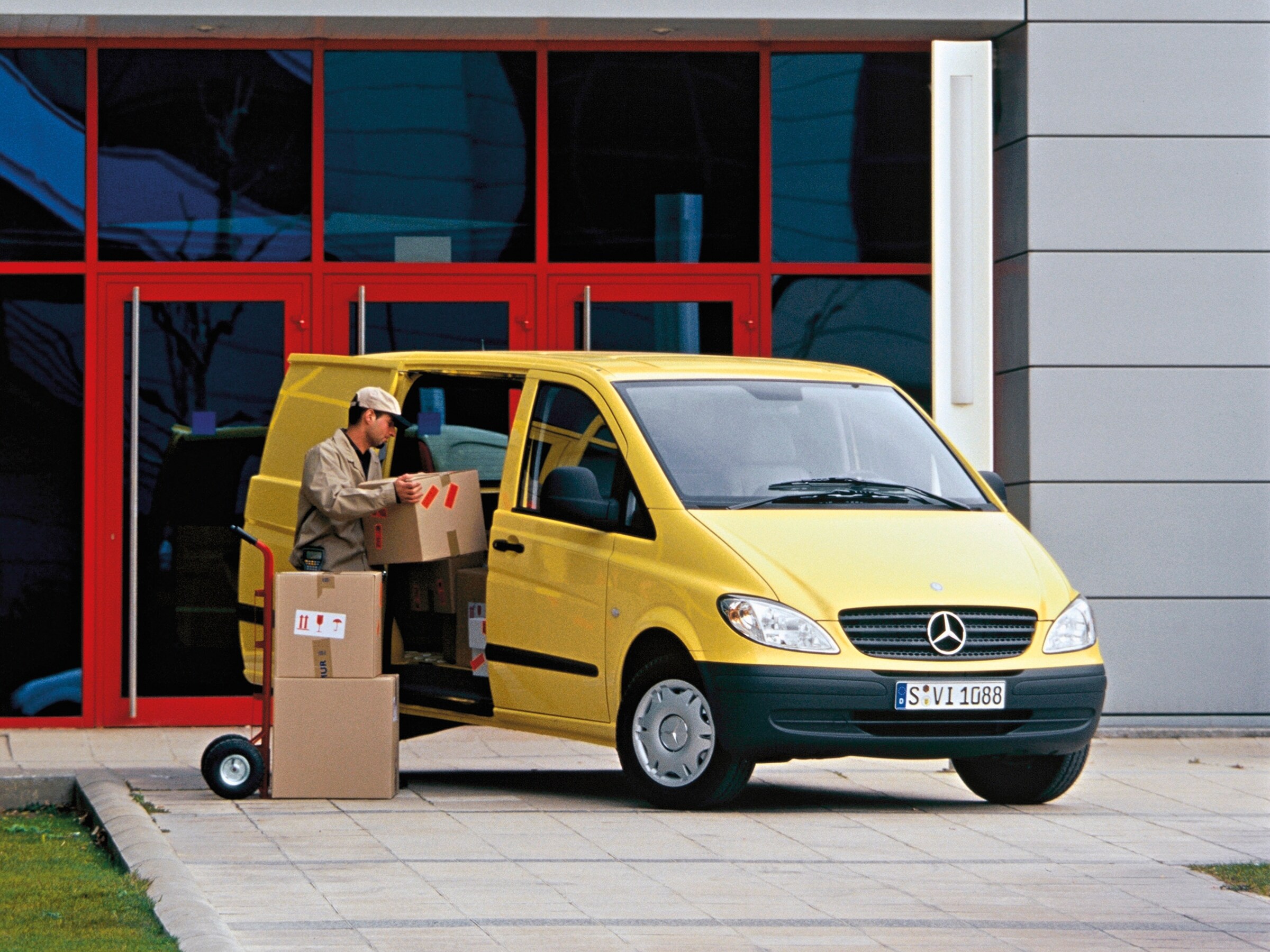 Un Mercedes-Benz Vito gialla parcheggiato davanti a un edificio, con un operaio che sta scaricando dei pacchi