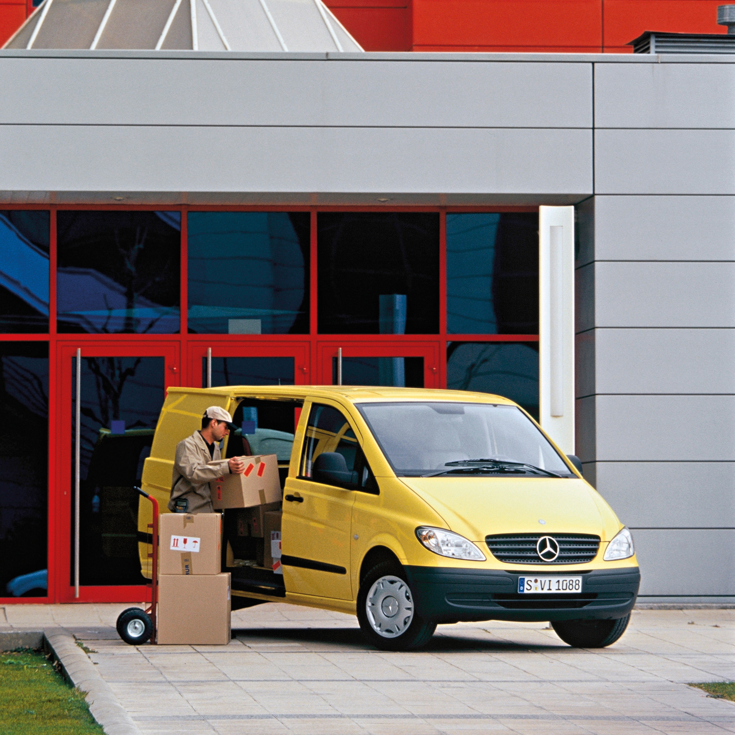 La seconda generazione dei Mercedes-Benz Vito | Storia dei veicoli commerciali | Mercedes-Benz Un Mercedes-Benz Vito gialla parcheggiato davanti a un edificio, con un operaio che sta scaricando dei pacchi