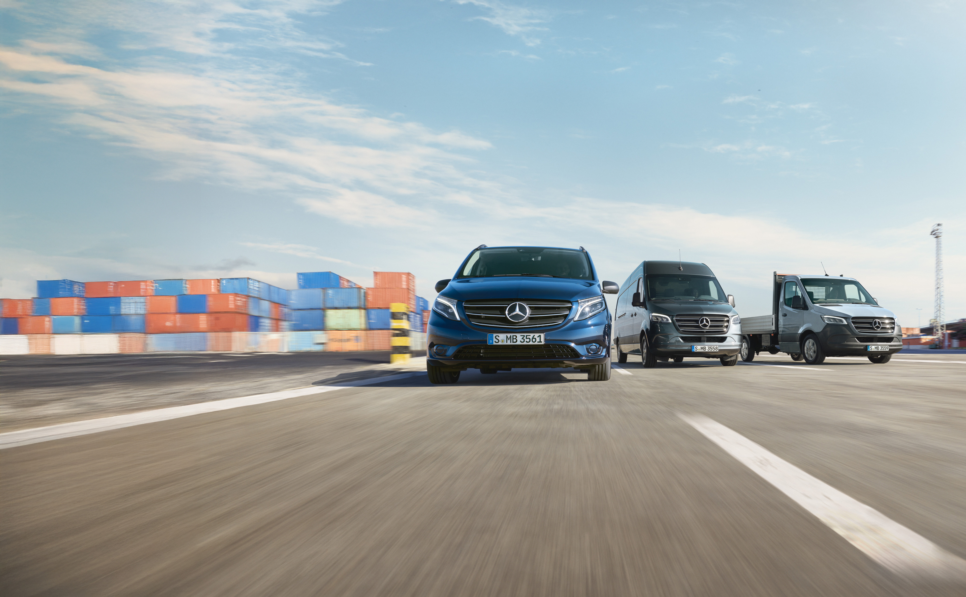 Mercedes-Benz | Assistenza veicoli commerciali | Service24h Una serie di veicoli commerciali Mercedes-Benz parcheggiati uno accanto all'altro su una strada accanto a un molo di spedizione.