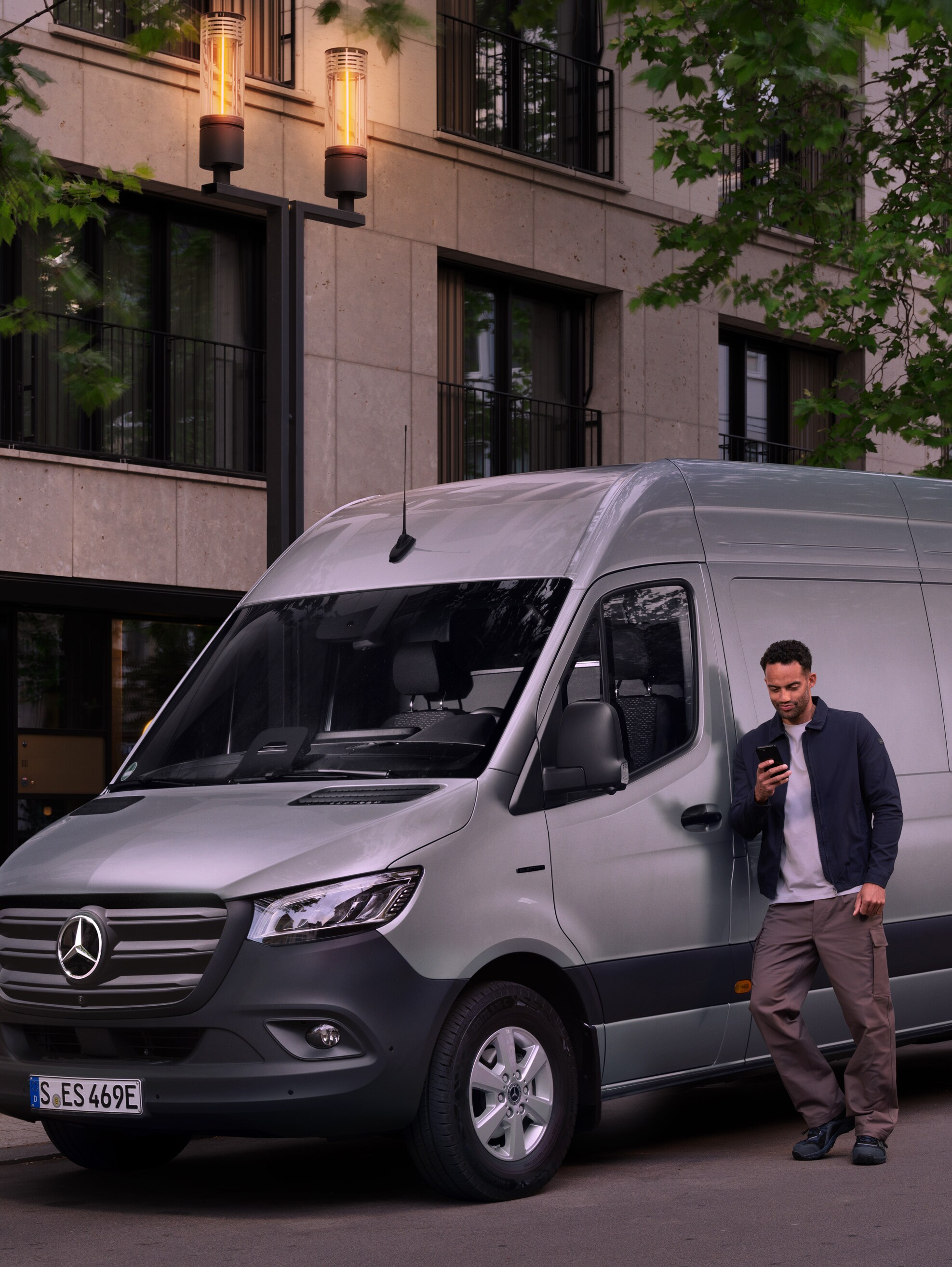 Un uomo appoggiato a un veicolo commerciale Mercedes-Benz Sprinter argento mentre utilizza il suo smartphone