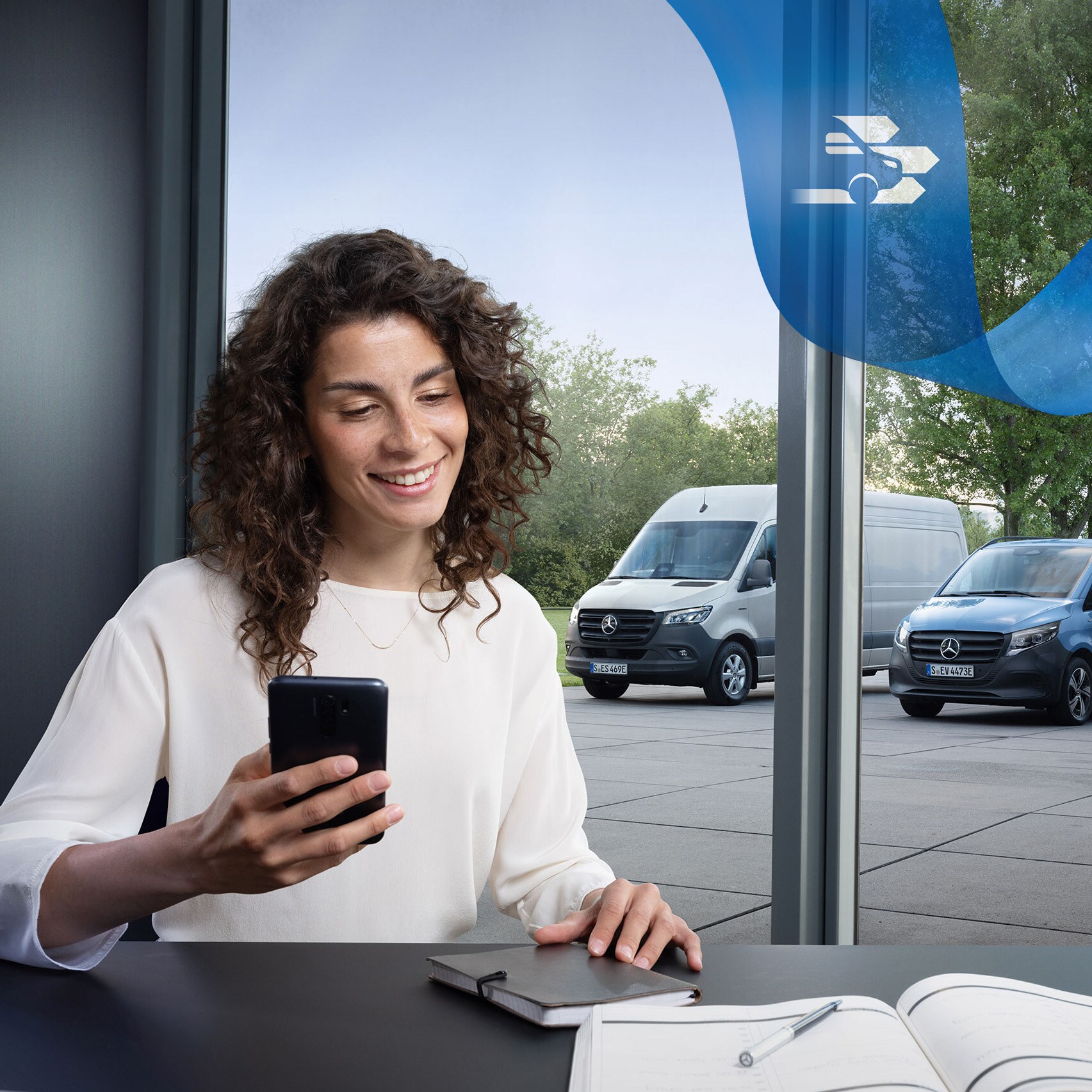 Una donna seduta alla scrivania con uno smartphone e una serie di veicoli commerciali Mercedes-Benz sullo sfondo.