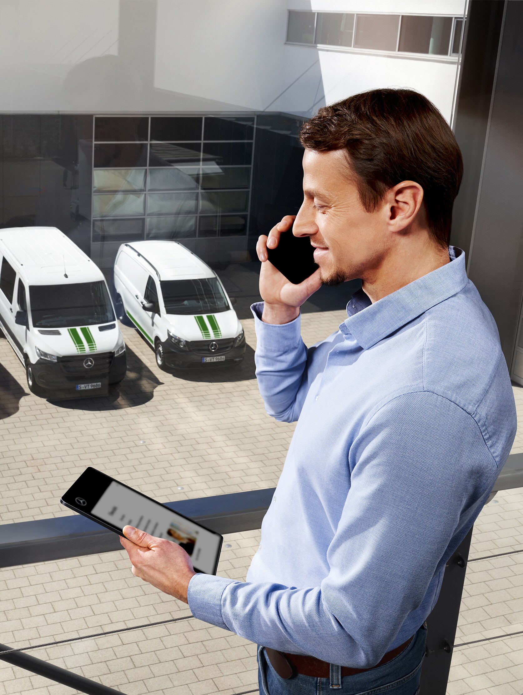 Un uomo al telefono con un tablet davanti a tre veicoli commerciali Mercedes-Benz