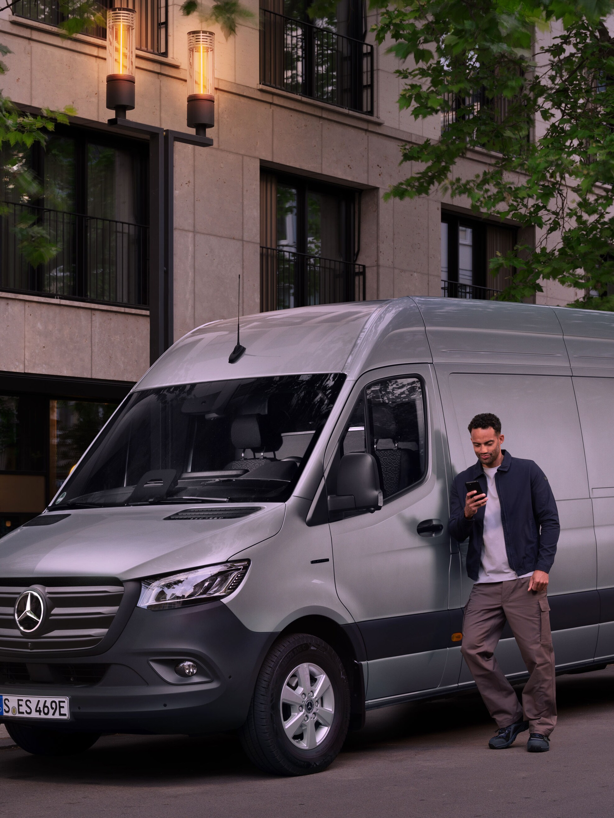 Un uomo appoggiato a un Mercedes-Benz Sprinter color argento mentre sta compilando un rapporto sui danni con il suo cellulare.