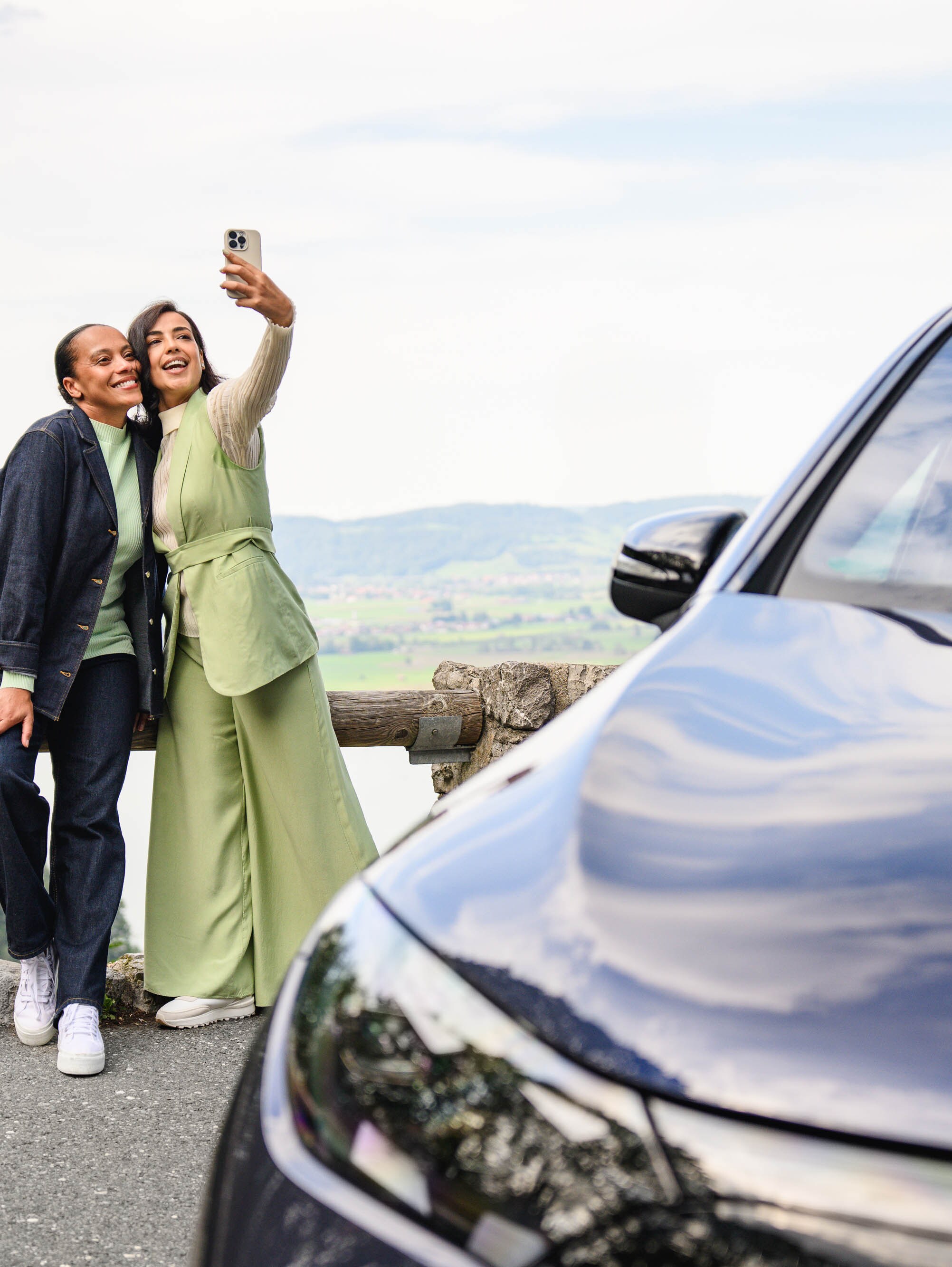 Due persone che scattano un selfie con il cellulare con lo sfondo di una montagna in lontananza mentre si trovano accanto a un veicolo Mercedes-Benz.