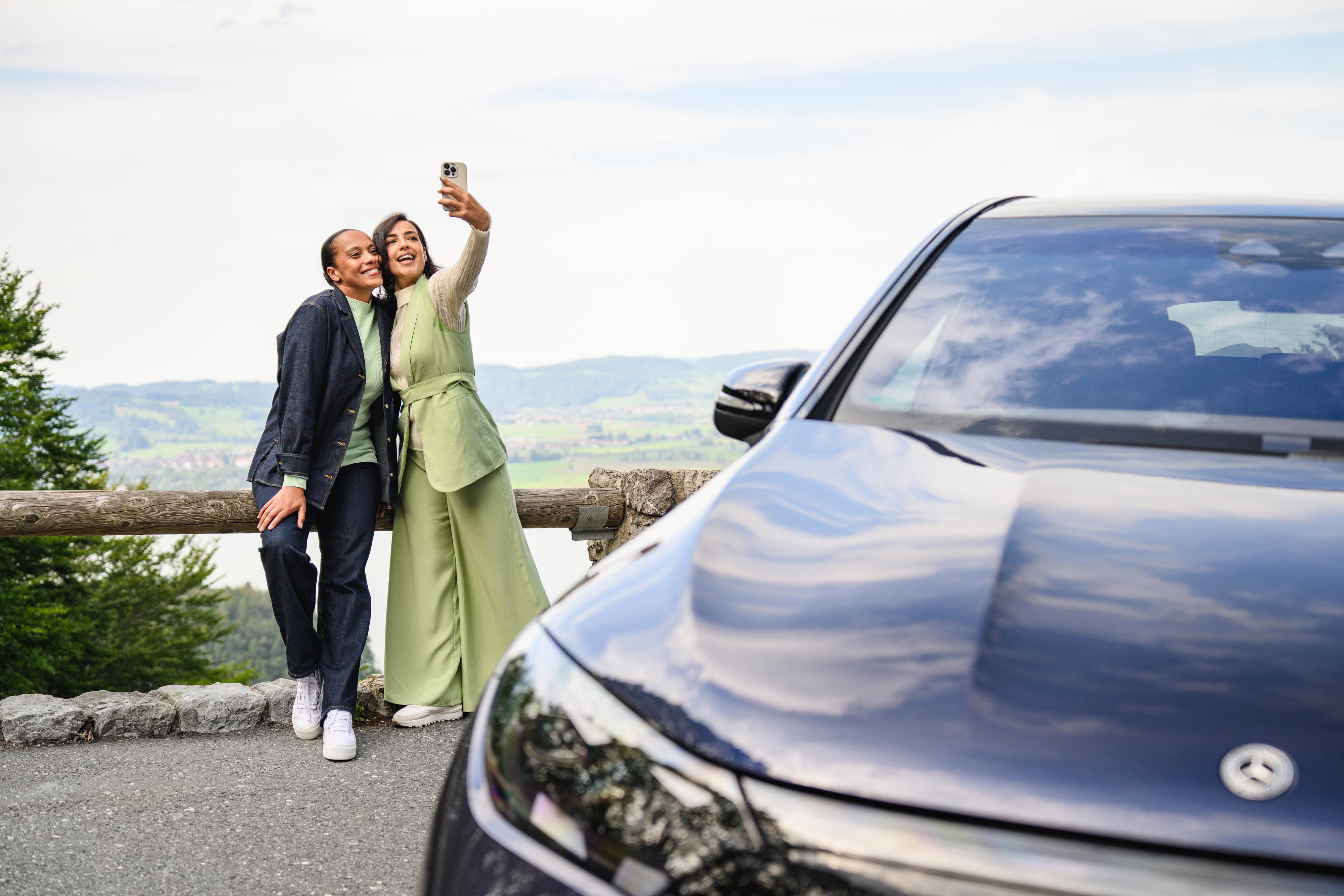 Noleggio a medio termine | Mercedes-Benz Rent Due persone che scattano un selfie con il cellulare con lo sfondo di una montagna in lontananza mentre si trovano accanto a un veicolo Mercedes-Benz.