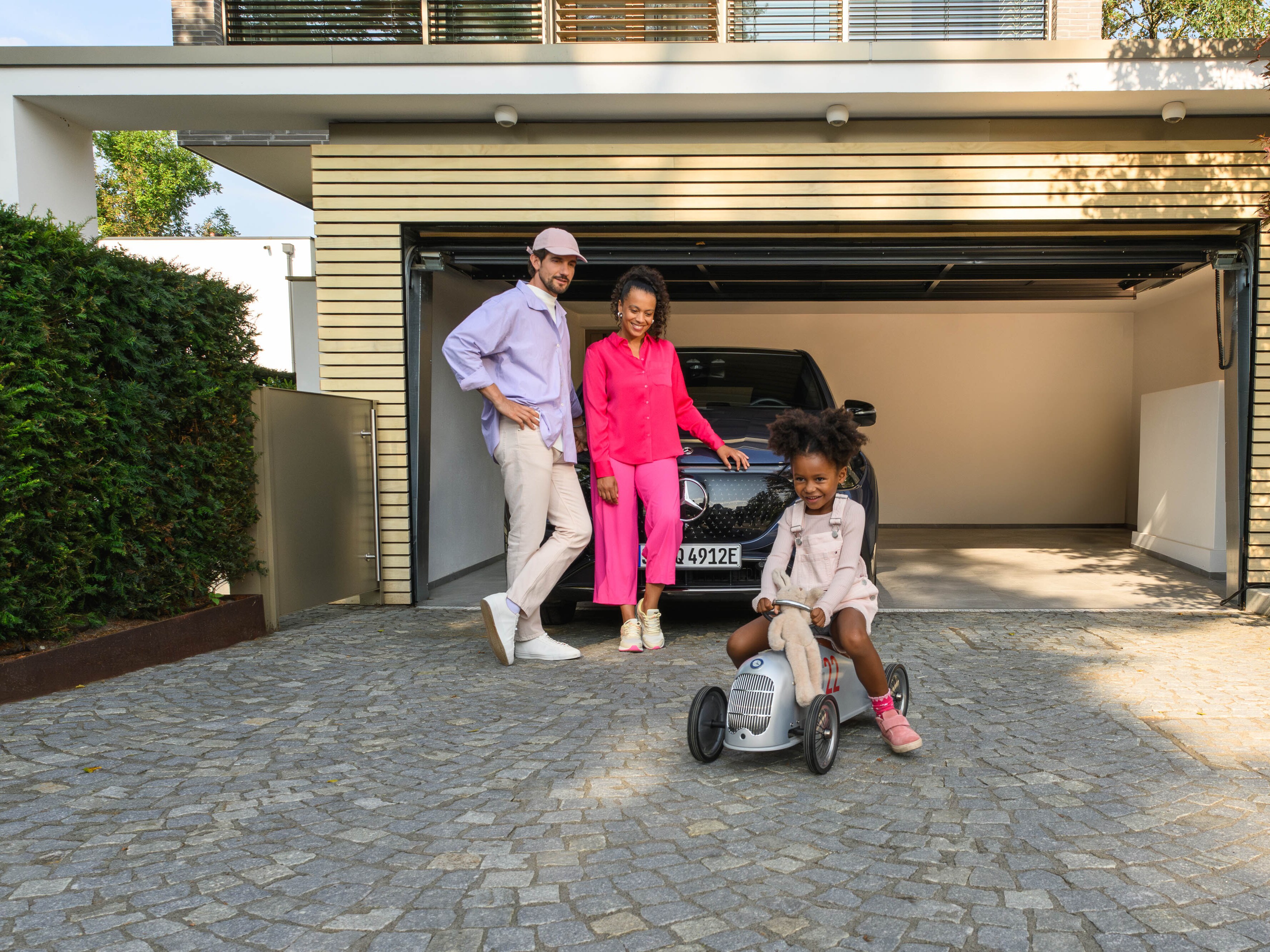 Due adulti appoggiati ad una Mercedes – Benz e una bambina che gioca con automobile giocattolo in un garage